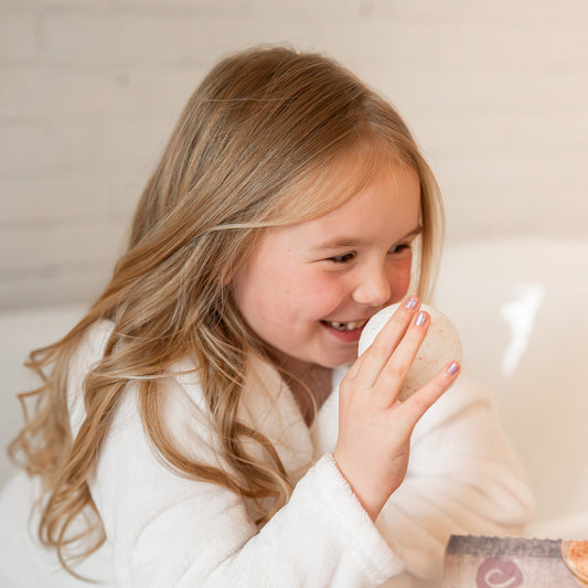 Young girl in a white robe smiling while holding a round, speckled Himalayan salt toning therapy bath bomb.