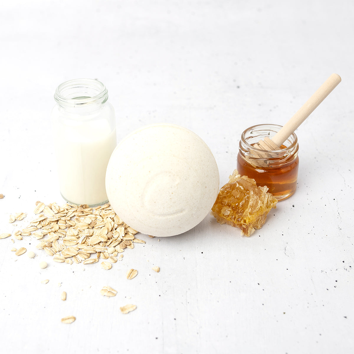 Cream-colored round bath bomb with a C-logo, surrounded by raw oats, a small jar of milk, and honey with a wooden dipper.