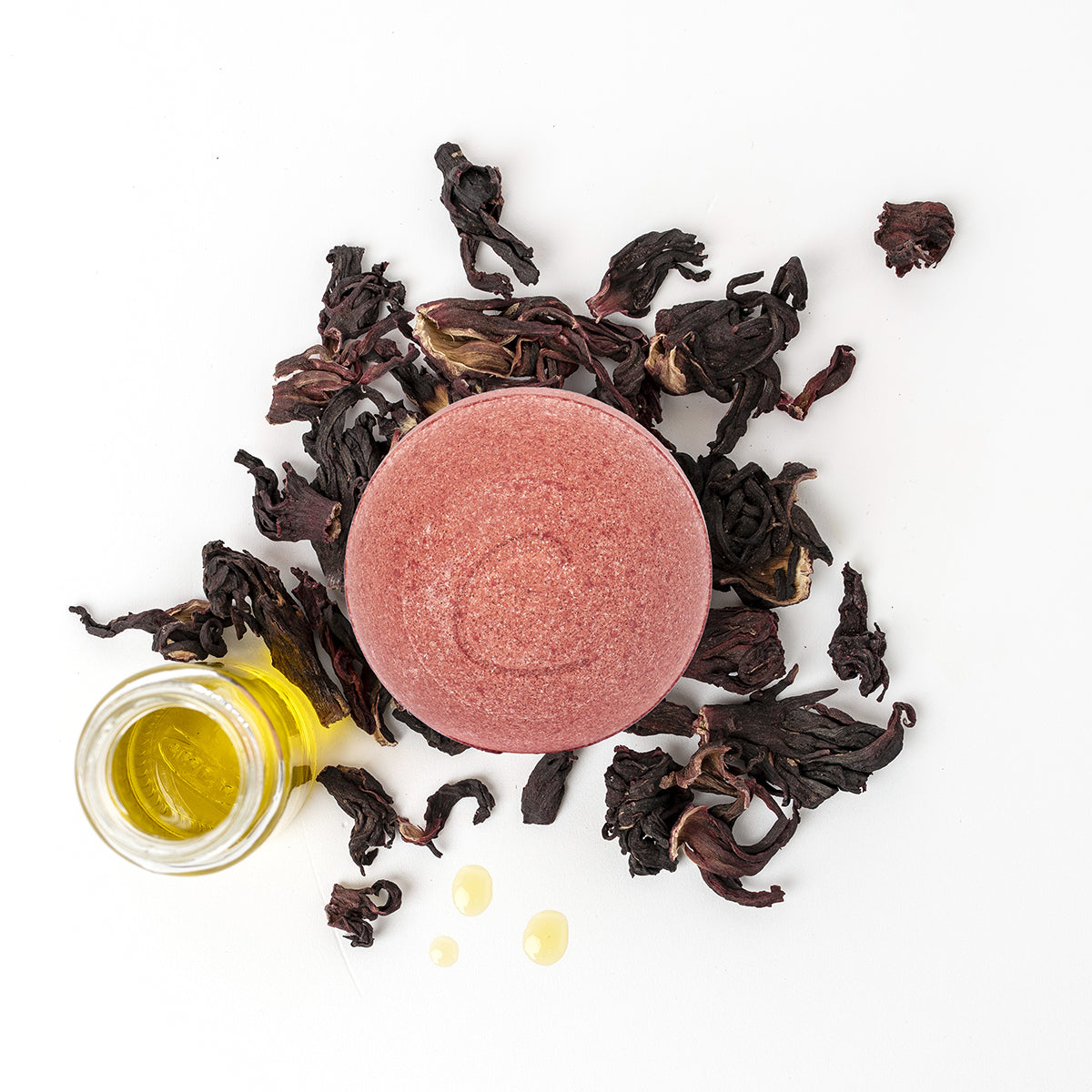Round pink bath bomb surrounded by dried hibiscus flowers and a small glass bottle of golden essential oil.