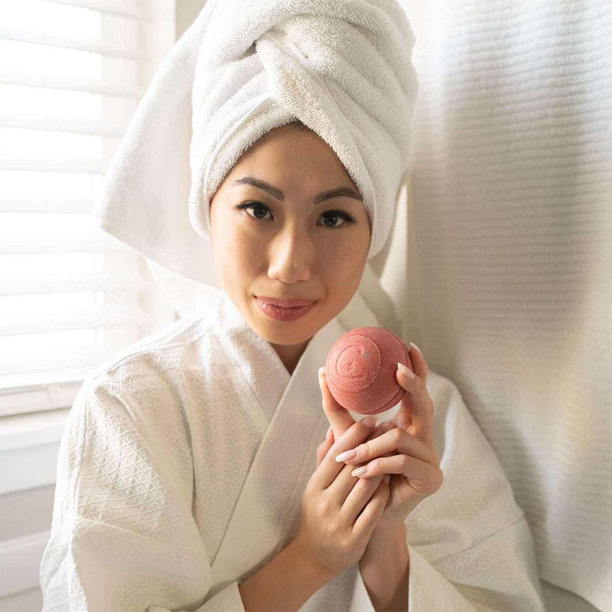 Woman in a white bathrobe and towel wrap holding a round, pink Hibiscus Passion Therapy bath bomb.
