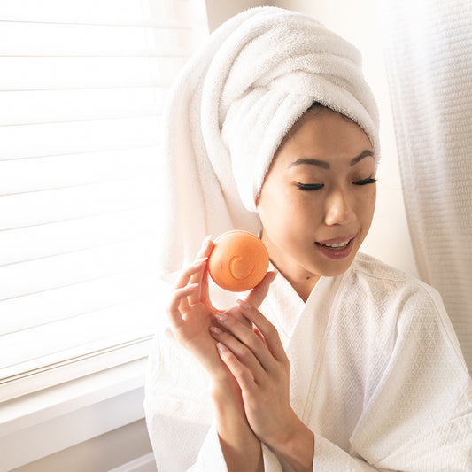 Woman in a white bathrobe and towel wrap holding a round, bright orange Kalahari Melon bath bomb.
